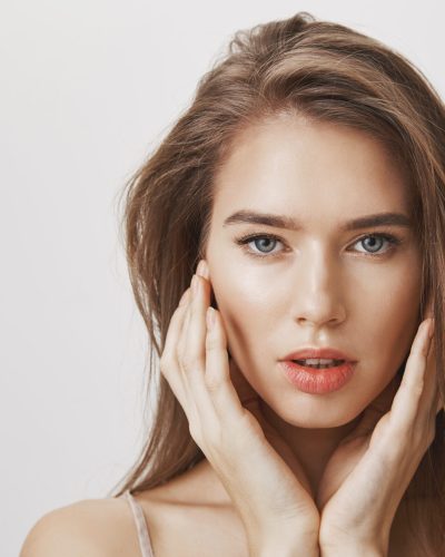 Studio shot of romantic attractive woman with natural makeup gently touching face and gazing at camera with focused confident expression, feeling relaxed and self-assured of her beauty over gray wall.